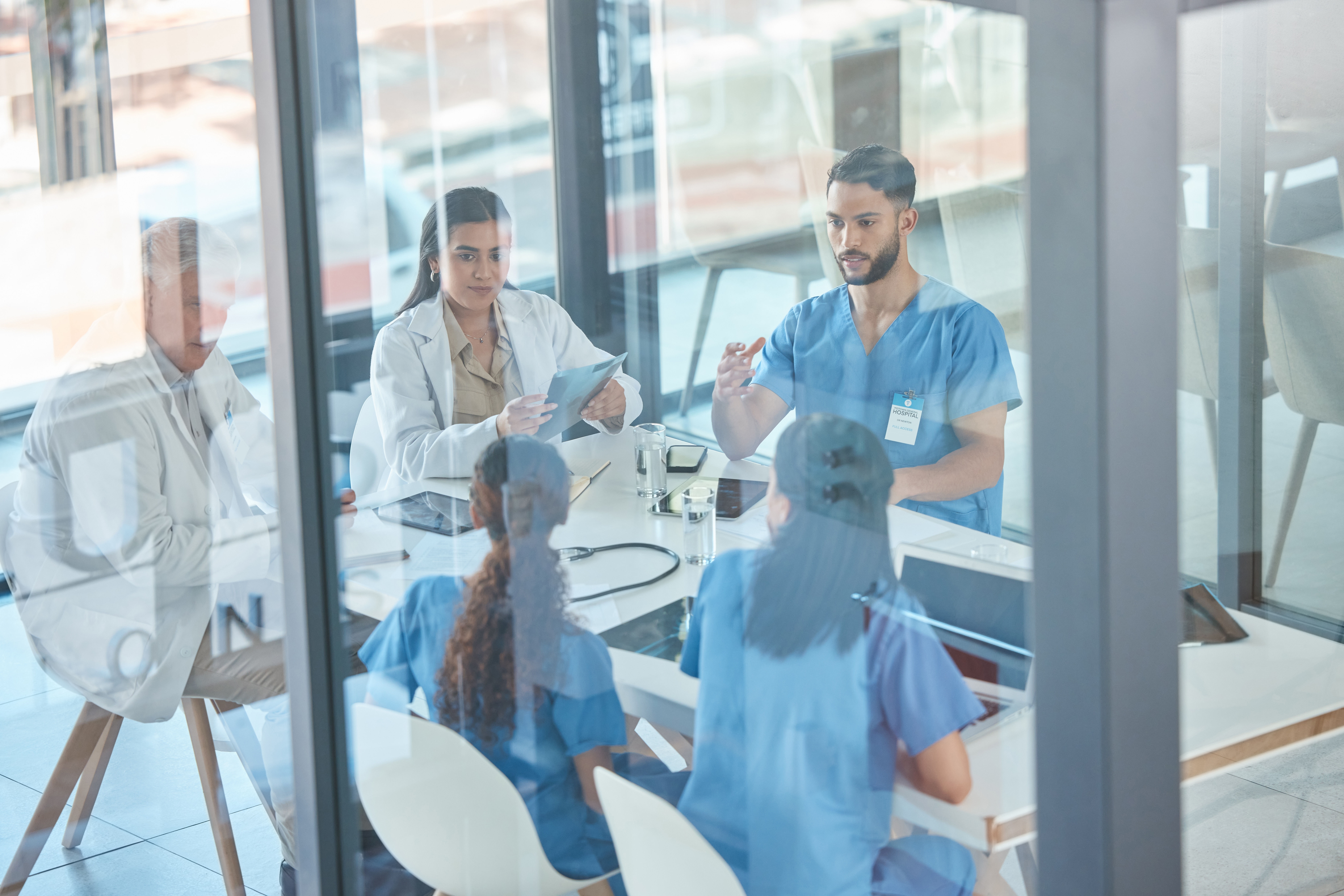 Group of healthcare professionals in scrubs and lab coats meeting around a conference table in a glass-walled office.