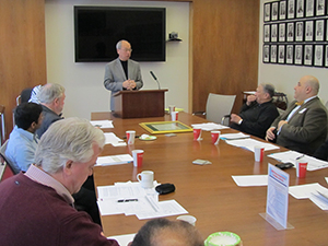 An image of members gathered around a table while a gentleman standing at a podium speaks to the members of IMG meeting in 2014